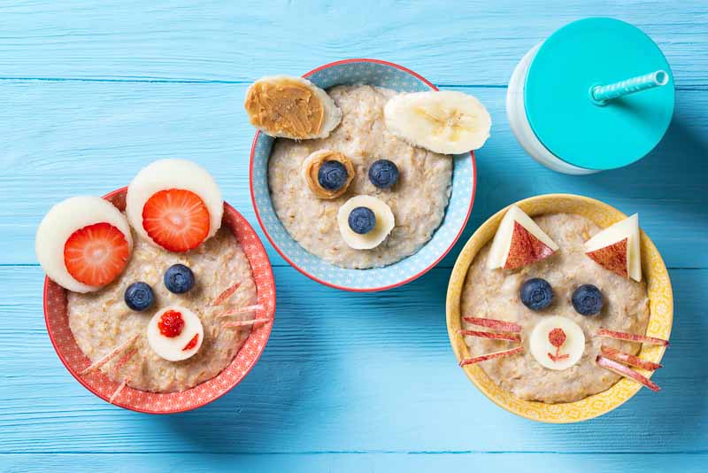 Funny bowls with oat porridge with cat, dog and mouse faces made of fruits and berries, food for kids idea, top view, blue wooden background